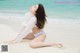 A woman in a white shirt and blue bikini sitting on the beach.