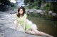 A woman in a green dress sitting on a rock by the water.